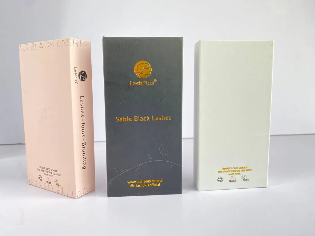 Three finished lash boxes standing closed: matte pink, black with a gold foil logo, and cream